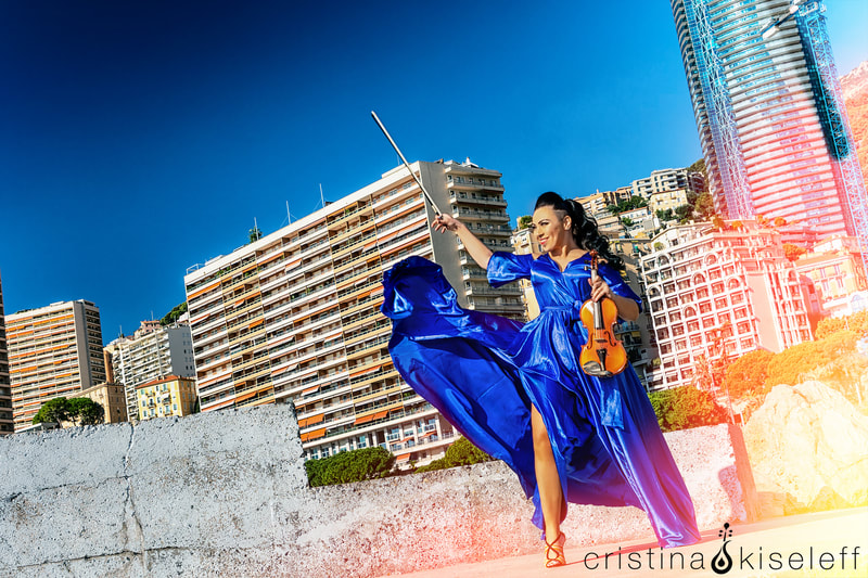 Cristina Kiseleff Electric Violinist plays the violin in Monaco waterfront with tall luxury buildings in a blue elegant dress